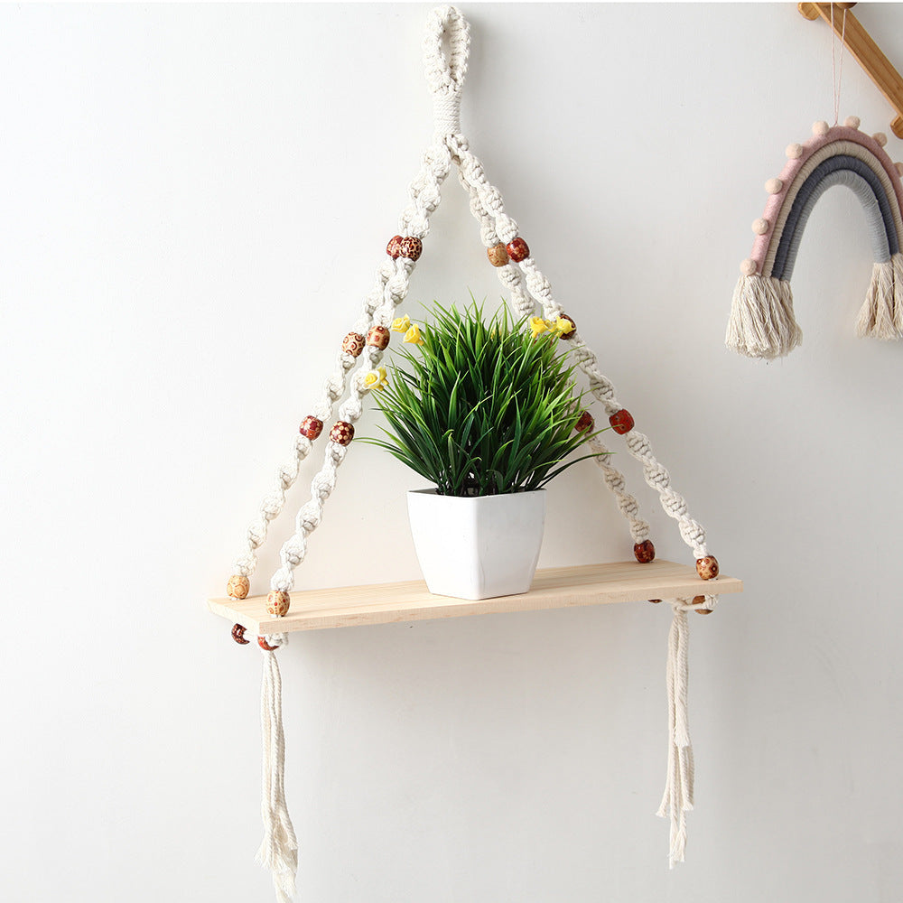 Macrame plant hanger with a wooden shelf holding a potted plant against a white wall.