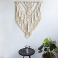 Macrame wall hanging on a white wall with a small table and plant in the foreground.