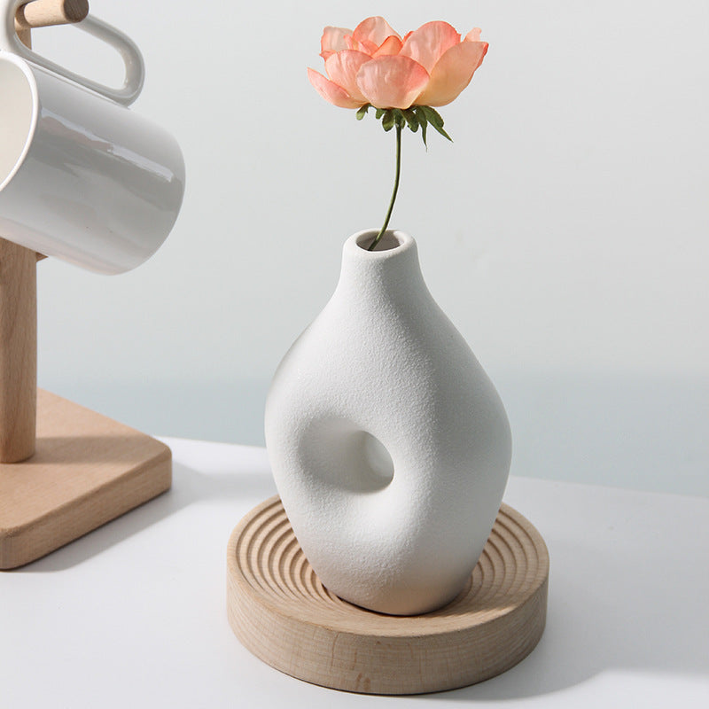 White textured vase with a flower on a wooden coaster, with a white mug and wooden stand in the background.