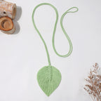 Green macrame leaf pendant necklace on a white background with wooden elements and dried plants.