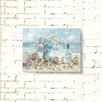 Framed artwork of a beach scene with flowers, shells, and a bottle on a white brick wall.