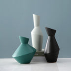 Three geometrically shaped vases in teal, white, and black on a light surface with a gray background. Velora Geometric Ceramic Vase.