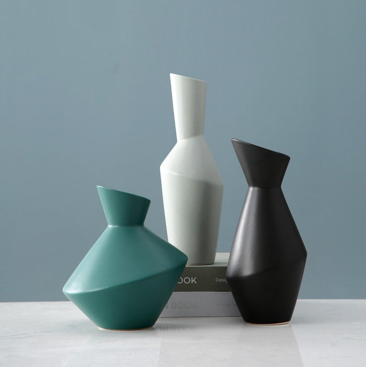 Three geometrically shaped vases in teal, white, and black on a light surface with a gray background. Velora Geometric Ceramic Vase.