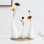 Three white vases with dried flowers on a woven mat against a white wall.